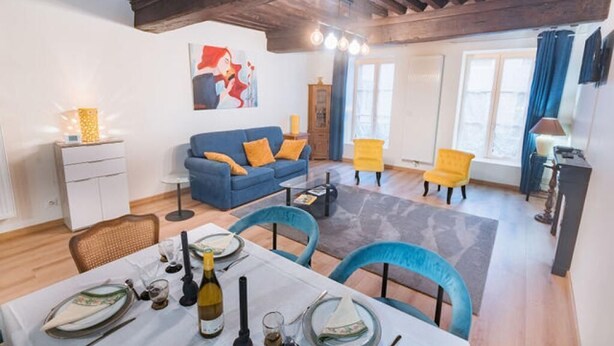 A living room with a blue sofa, yellow armchair, dining table set with plates and wine glasses, and a painting on the wall. - Charming townhouse with a covered and furnished interior patio (Tournus)