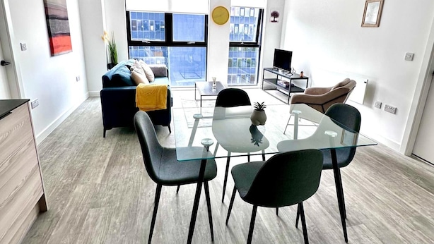 Una sala de estar moderna con una mesa de comedor de vidrio, sillas negras, un sofá con cojines y un televisor sobre un mueble bajo.
