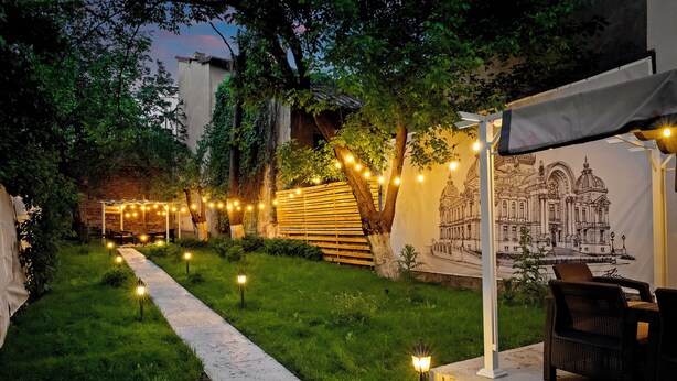 A garden pathway with string lights, a pergola, and a mural of a building.