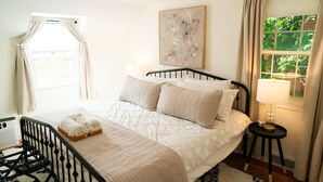 Un dormitorio con cama, mesita de noche con lámpara, ventana con cortinas y un cuadro abstracto colgado en la pared.