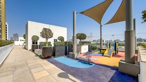 A playground with colorful play equipment, surrounded by planters and a white building.