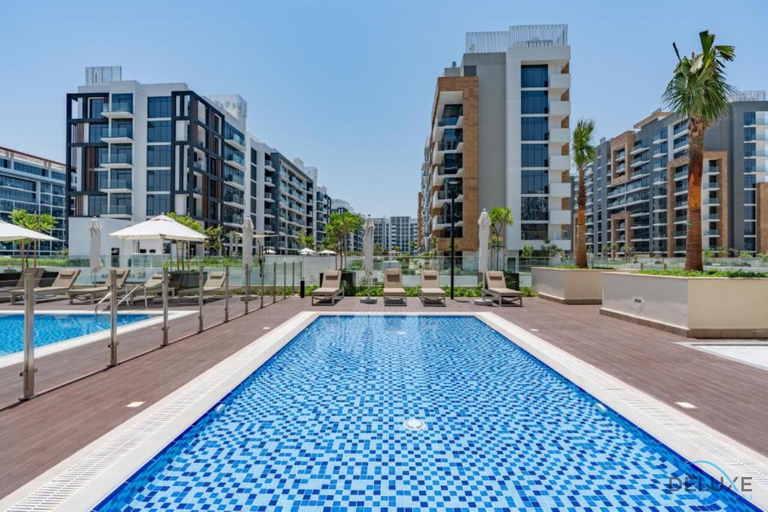 Una piscina con sillones de descanso y sombrillas, rodeada de modernos edificios de apartamentos.