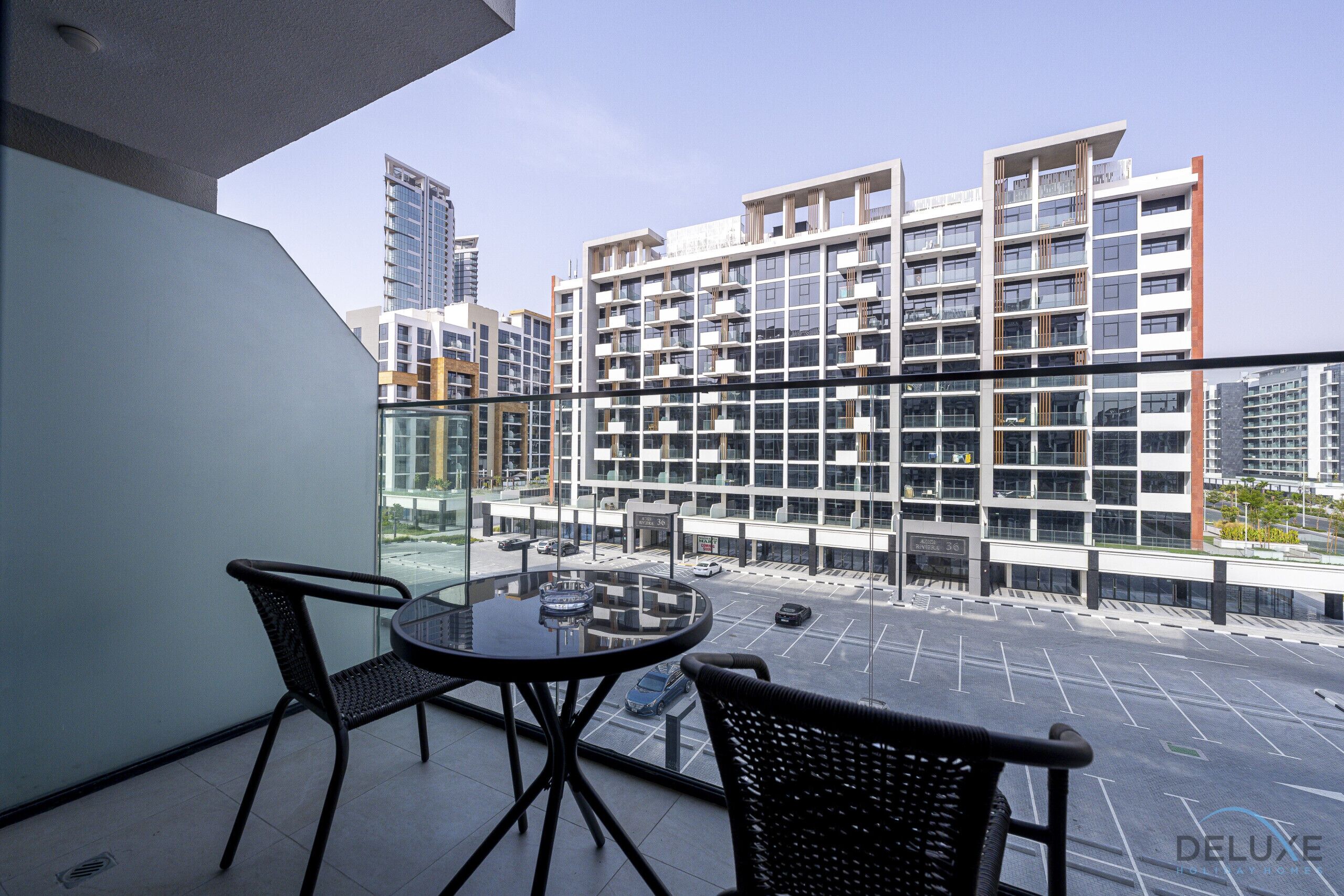Un balcón con dos sillas y una mesa, con vistas a un estacionamiento y edificios modernos.