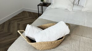 Een net opgemaakt bed met witte lakens, een geweven mand met een witte handdoek, en een klein nachtkastje met een lamp en een plant.