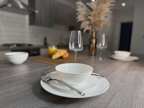 A modern kitchen with a dining setup featuring a white bowl and plate on a wooden table, a vase with dried flowers, and a bottle of orange juice.