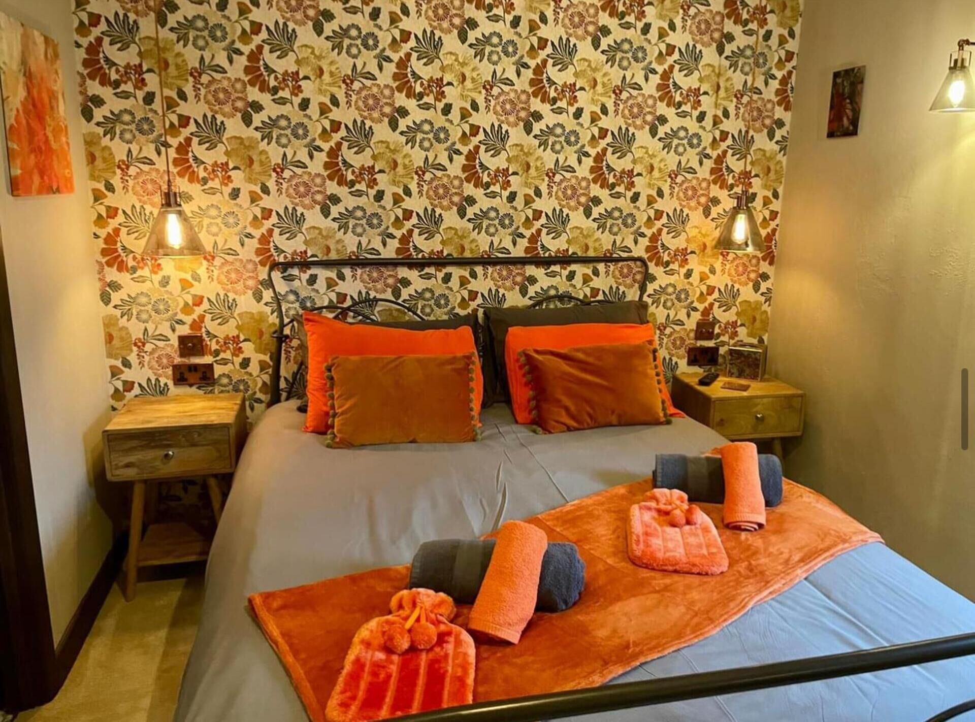 Un dormitorio con papel tapiz floral, estructura de cama metálica, dos mesitas de noche y un tocador.