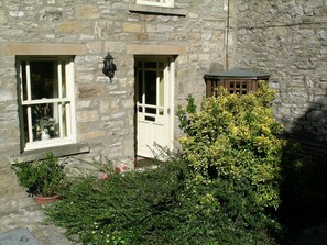A stone building with two windows and a door, surrounded by greenery.