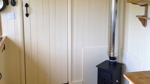 A room with a wood-burning stove, a door with a black handle, a white wall, and a wooden floor. - Beeches Shepherd Hut (Harleston)