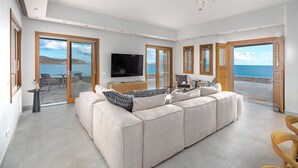 Ein geräumiges Wohnzimmer mit einem großen, beigefarbenen Ecksofa, einem Flachbildfernseher und Meerblick durch große Glasüren.