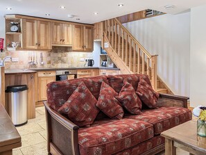 Uma sala de estar aconchegante com um sofá vermelho estampado, escada de madeira e uma área de cozinha com armários de madeira e painel de azulejos.