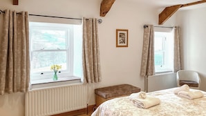 A bedroom with a large bed, a window with curtains, a radiator, a wooden floor, and a small bench. - 1 Swallowholm Cottages (Arkengarthdale)