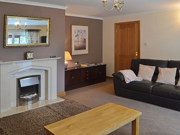 A living room with a fireplace, a black leather sofa, a wooden coffee table, a dark wooden cabinet, a framed picture on the wall, and a door. - 4 bedroom accommodation in Red Row near Amble (Red Row near Amble)