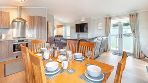 A modern kitchen with stainless steel appliances, a dining area with a wooden table set for four, and a living area with a sofa and TV.