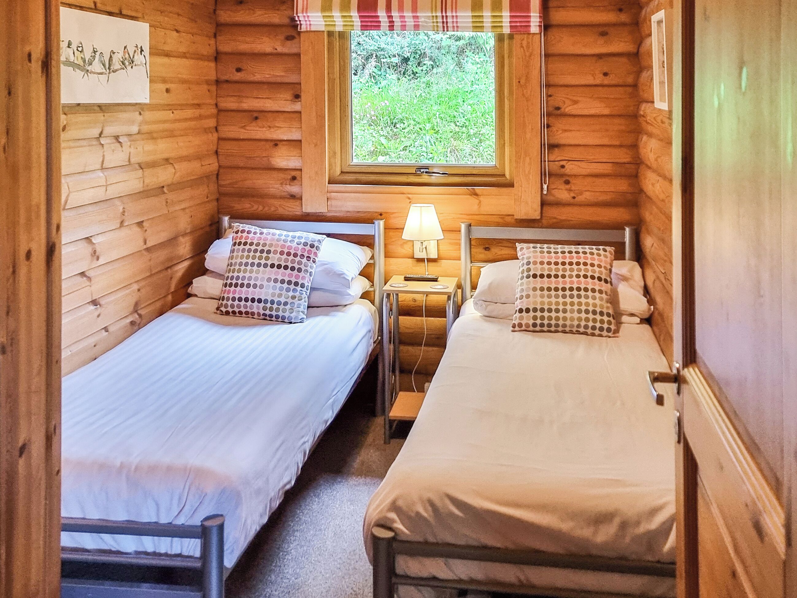 Une cabane avec deux lits en bois, une fenêtre donnant sur la verdure, une lampe fixée au mur et un tableau.