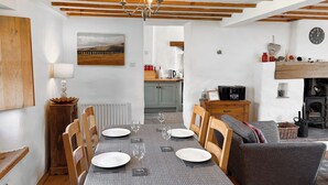 A dining area with a table set for four, wooden chairs, a chandelier, a painting on the wall, and a kitchen in the background.