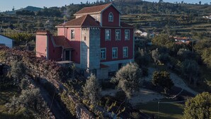 Une grande maison à plusieurs étages, à la façade rouge et grise, entourée de verdure et de collines.