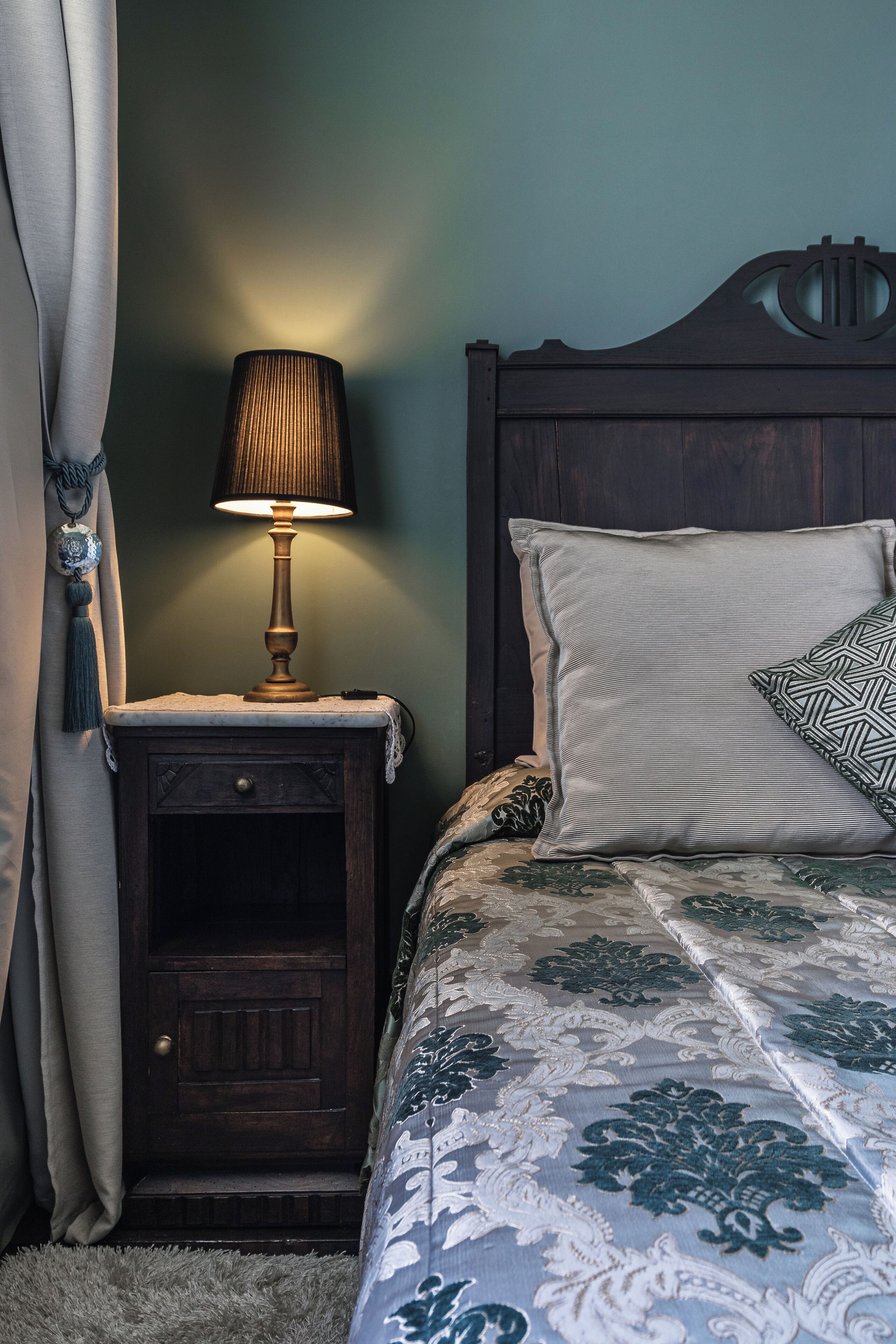 Un dormitorio con una mesita de noche de madera, una lámpara, una cama con ropa de cama estampada y un cabecero.