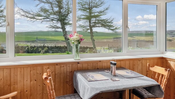 Een gezellig hoekje met een houten tafel en stoelen, een vaas met bloemen, en uitzicht op een groen veld door grote ramen.