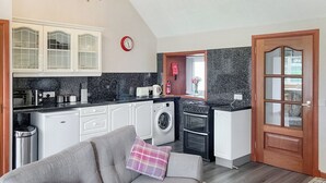 Een compacte keuken met witte kasten, een inbouwoven, een wasmachine en een eethoek met een grijze bank en een ruitkussen.