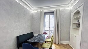 A dining area with a marble table, two chairs, a bench, and a view through a window with curtains.
