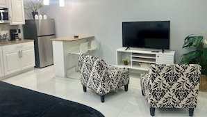 Ein modernes Wohnzimmer mit Flachbildfernseher, zwei gemusterten Sesseln, einer Küchenzeile mit weißen Schränken und Kühlschrank sowie einem Esstisch mit Stühlen.