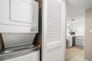 A laundry area with a front-loading washer and dryer, a white door with horizontal slats, and a kitchen in the background.