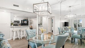 Un interior moderno con una mesa de comedor de vidrio, taburetes de bar y una cocina con armarios y electrodomésticos blancos.
