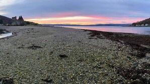 A pebbly beach with a castle in the background and a sunset sky. - A perfect location for a family island break by the seaside. (Lochranza)