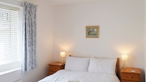 A bedroom with a bed, two bedside tables, a nightstand, a window with blinds, and a framed picture on the wall.