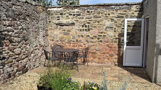A stone wall with a wooden trellis, a table with chairs, and potted plants in a courtyard.