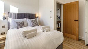 A neatly made bed with white linens and two rolled towels, a wooden door, and a small bedside table with a vase.