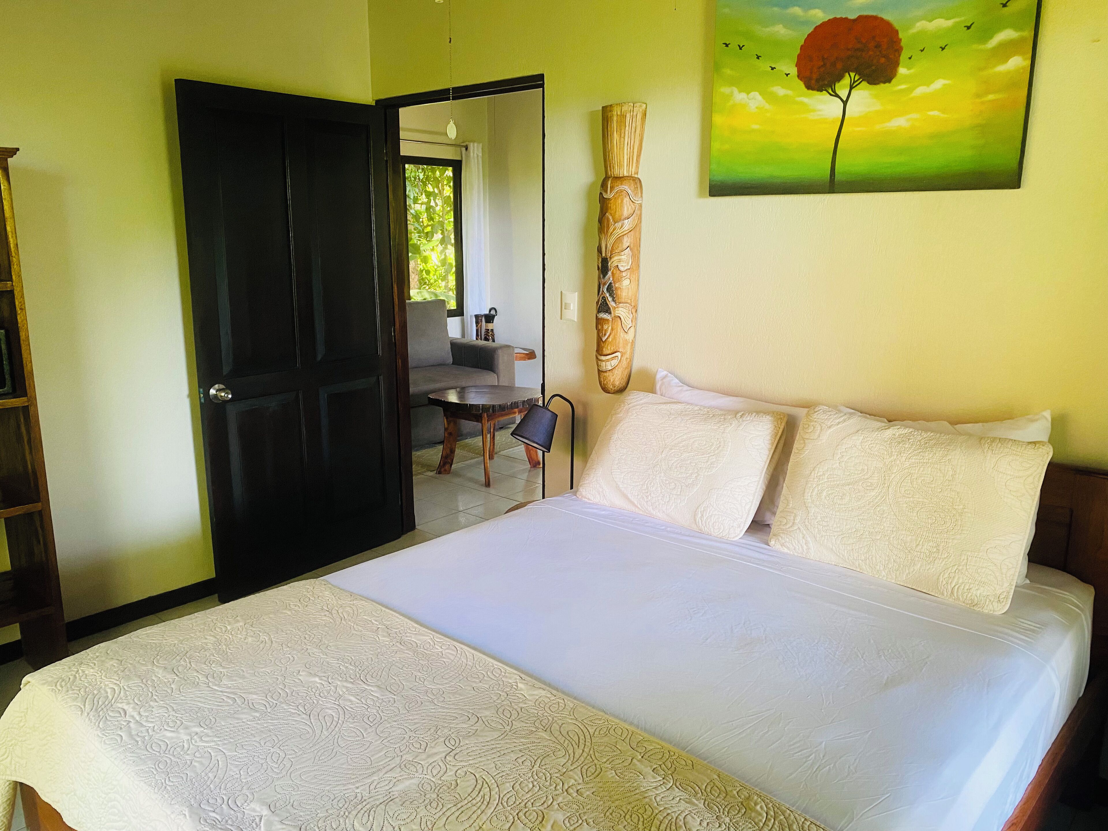 Un dormitorio con cama, almohadas, una puerta de madera, un cuadro y vistas al exterior.