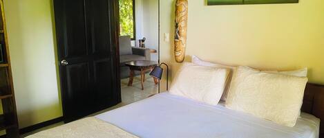 A bedroom with a bed, pillows, a wooden door, a painting, and a view of the outdoors.