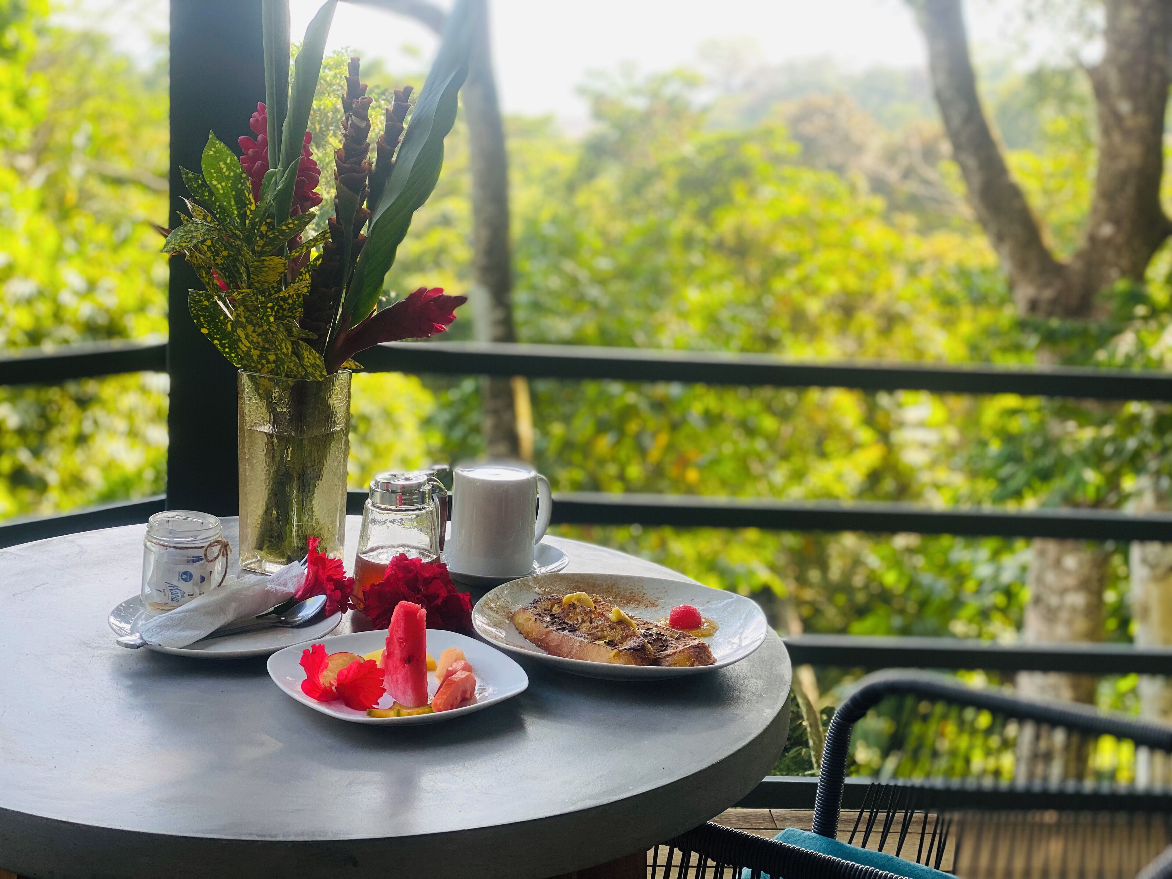 A table with a glass of water, a mug, a plate with food, and a vase with flowers on a balcony with a view of greenery.