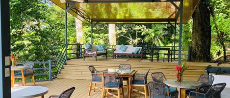Un patio al aire libre con suelo de madera, sillas de mimbre, una mesa y una zona de estar cubierta con cojines.