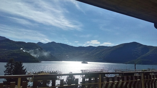 Ein See mit Bergen im Hintergrund, ein Boot auf dem Wasser und ein Gebäude mit Balkon im Vordergrund.