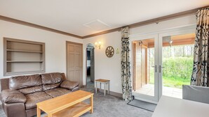 A living room with a brown leather sofa, a wooden coffee table, and a white dining table. There is a door leading to a balcony with a view of greenery. - A peaceful location for a relaxing break with excellent fishing to enjoy. (Marston)