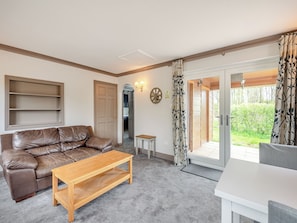 A living room with a brown leather sofa, a wooden coffee table, and a white dining table. There is a door leading to a balcony with a view of greenery. - A peaceful location for a relaxing break with excellent fishing to enjoy. (Marston)