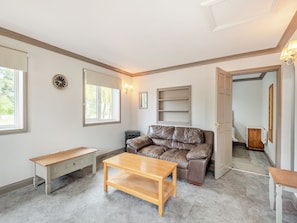 A room with a brown leather sofa, a wooden coffee table, a bench, a wall clock, a window with a view, and a door leading to another room. - A peaceful location for a relaxing break with excellent fishing to enjoy. (Marston)