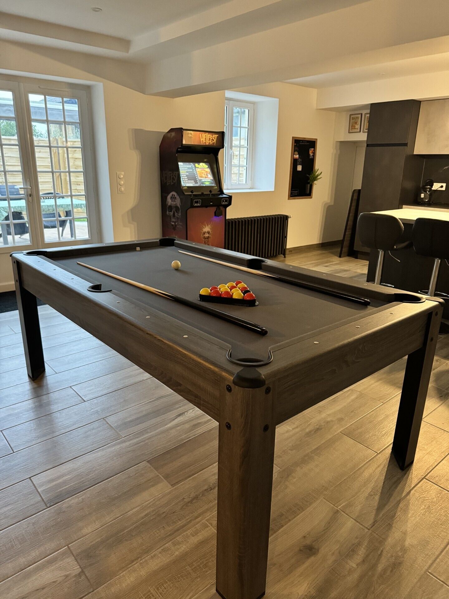 A pool table with a cue stick and balls, a pool table in a room with wooden flooring, a window, and a vending machine.