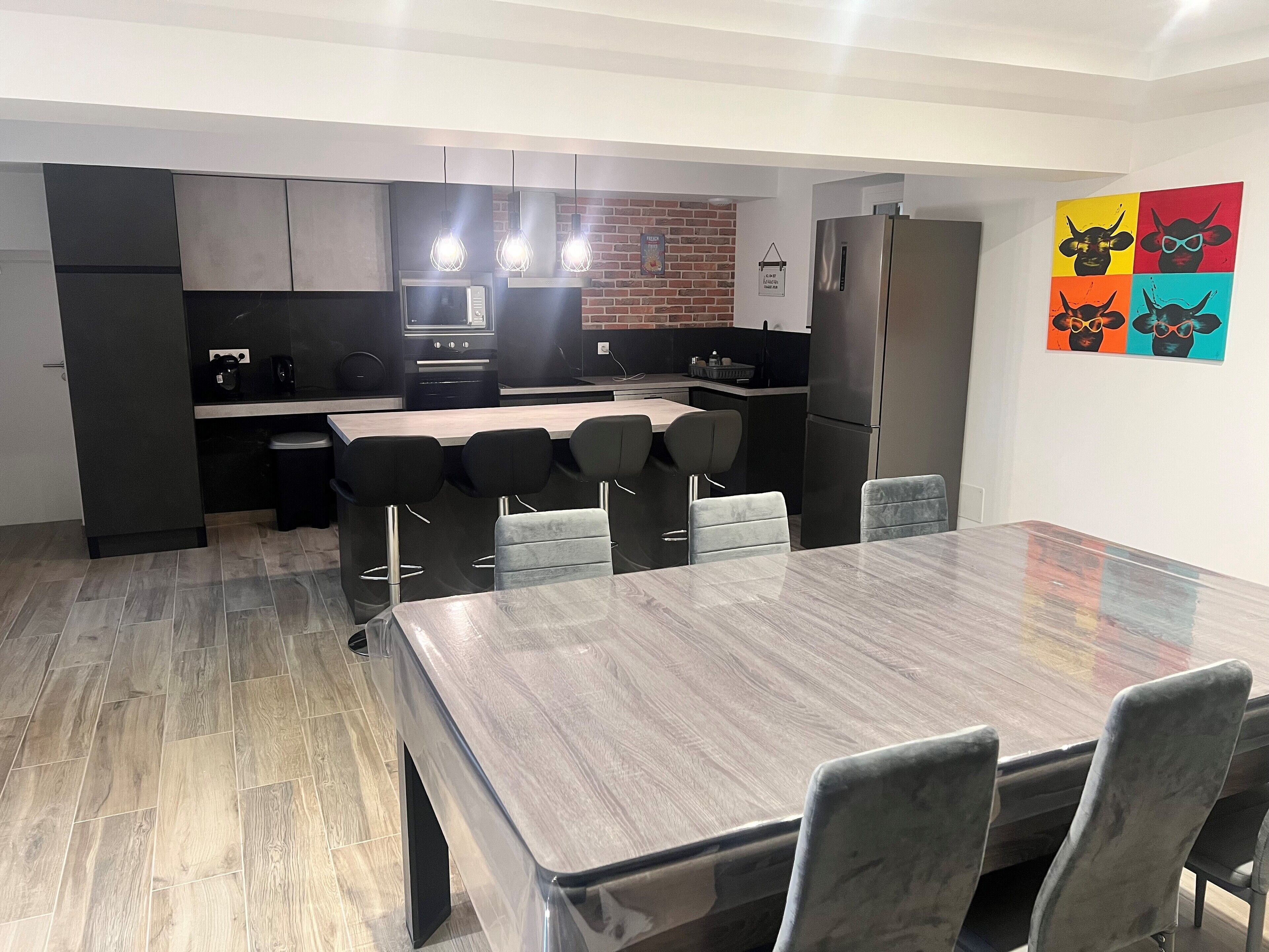 Une cuisine moderne avec un coin repas, comprenant une grande table en bois et des tabourets de bar, un réfrigérateur et un mur de fond en briques.