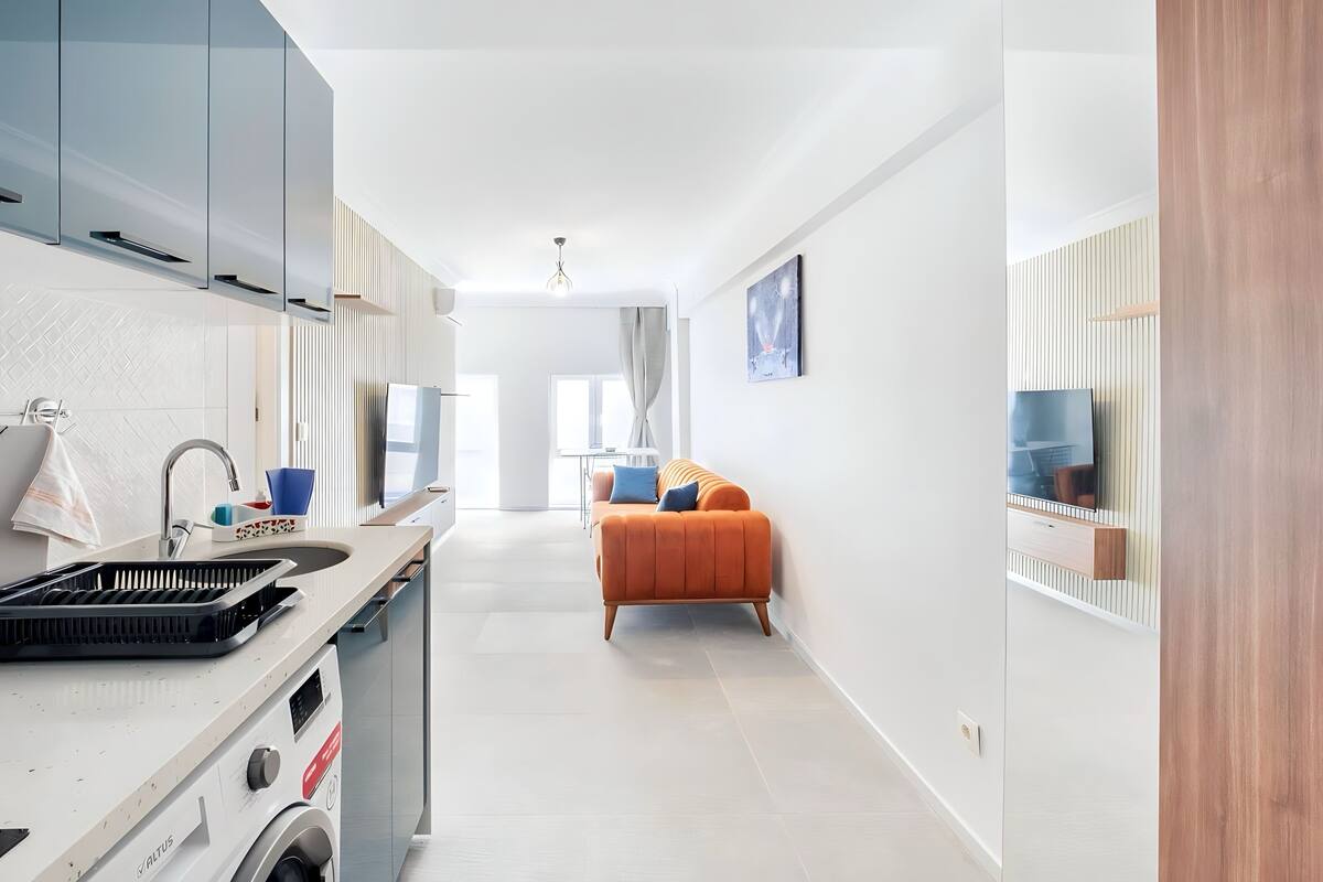 Una cocina moderna con lavadora incorporada, un comedor con sofá y un amplio ventanal que proporciona luz natural.