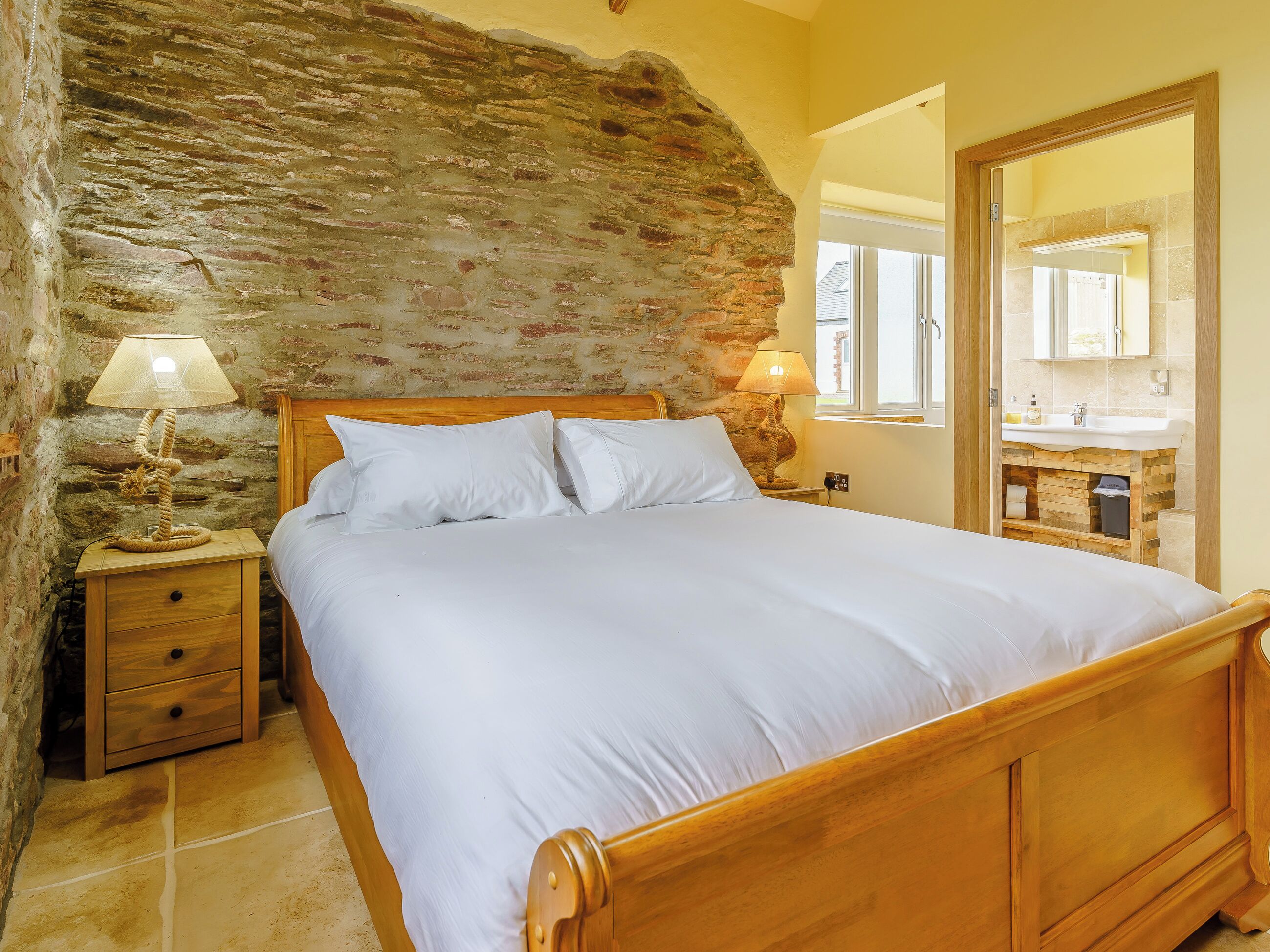 Ein Schlafzimmer mit Steinwand, einem hölzernen Bett, einem Nachttisch mit Lampe und einem durchgehenden Türspalt sichtbaren Badezimmer.