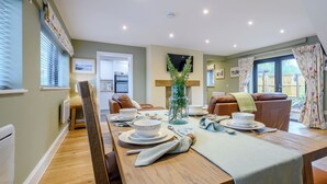 A dining area with a wooden table set for four, a kitchen in the background, and a view of the outdoors through a large window.