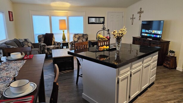A modern kitchen with a central island, dining area with a table and chairs, a living room with a sofa and armchairs, and a TV on the wall. - Private 10-Acre Retreat Near JBLM {Fast Wi-Fi} with Adjoining RV Pad (Yelm)