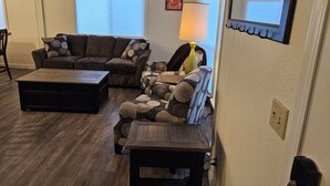 A living room with a dark brown sofa, a patterned armchair, and a wooden coffee table. There is a lamp on a side table, a painting on the wall, and a window with blinds. - Private 10-Acre Retreat Near JBLM {Fast Wi-Fi} with Adjoining RV Pad (Yelm)