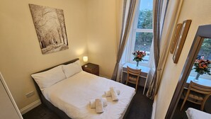 Una cama bien hecha con sábanas y toallas blancas, una mesita de noche con una lámpara, un escritorio pequeño con una silla y una ventana con cortinas.