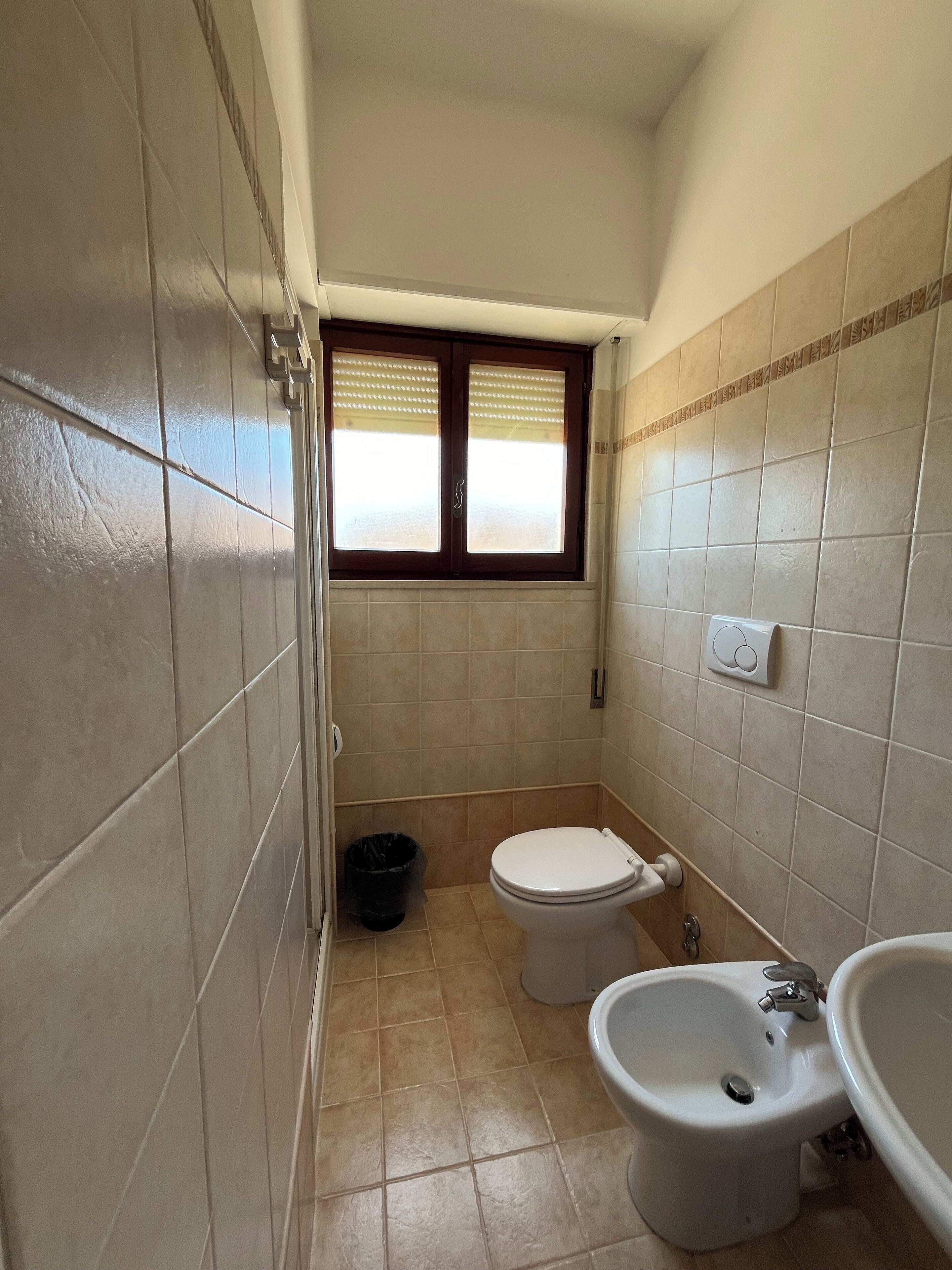A small bathroom with a toilet, bidet, and sink. There is a window with blinds, a trash bin, and tiled walls.