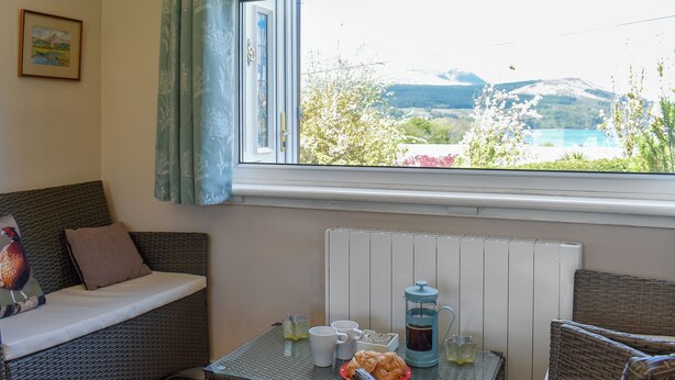 A cozy room with a wicker sofa, a small table with a coffee maker, and a view of a lake and mountains.