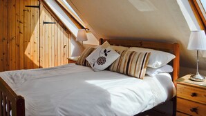 A bedroom with a sloped ceiling, wooden walls, a bed with white bedding, striped pillows, and a bedside table with a lamp.